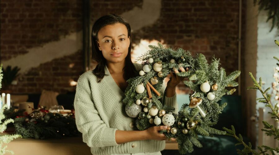 A Woman in Gray Sweater Holding a Christmas Wreath with Beautiful Design while Looking at the Camera