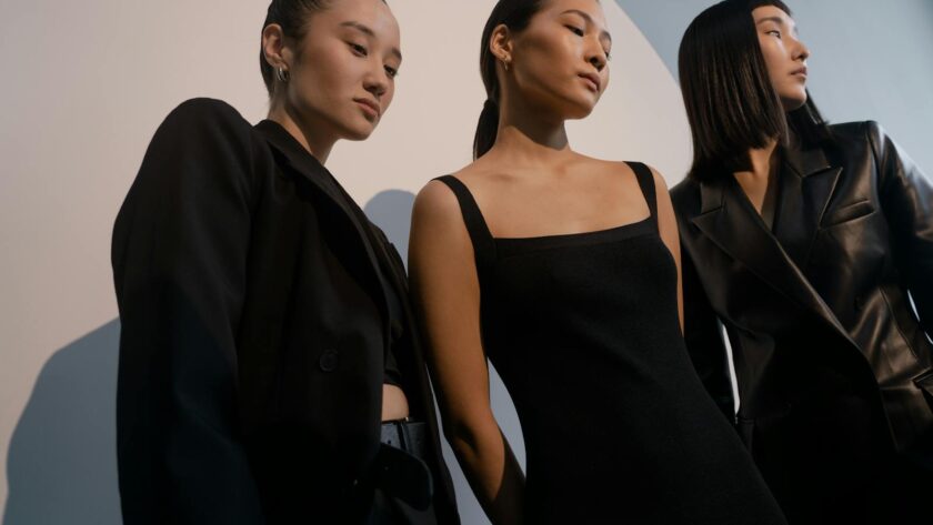 Three stylish women posing in chic black outfits against a minimalist backdrop.