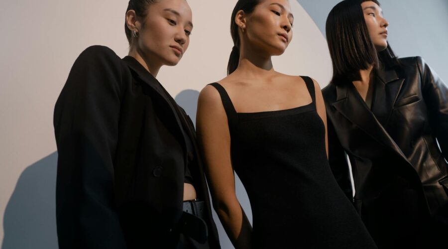 Three stylish women posing in chic black outfits against a minimalist backdrop.